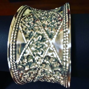 May include: A silver bangle bracelet with an intricate floral design. The bracelet is wide and has a beaded edge.