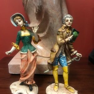 May include: A pair of vintage porcelain figurines depicting a man and a woman in 18th-century attire. The woman is wearing a green dress with a pink skirt and a blue hat. The man is wearing a brown jacket with blue pants and a yellow hat. Both figurines are standing on white bases.
