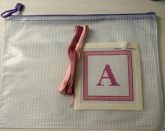 Puede incluir: Una bolsa de malla plástica transparente con cremallera que contiene un lienzo cuadrado de punto de aguja con una letra A rosa dentro de un borde rosa y melocotón, junto a dos madejas de hilo de bordar en tonos rosa oscuro y rosa claro.
