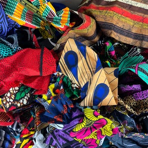 May include: A vibrant assortment of colorful African fabric scraps. The textiles feature bold patterns in shades of blue, yellow, green, red, and purple. The fabrics are layered and bunched together, showcasing a variety of designs and textures.