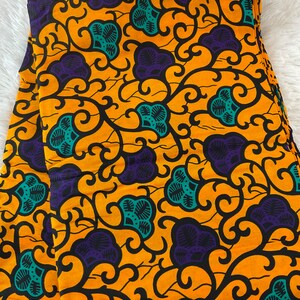 May include: Folded fabric with a vibrant floral pattern. The design features large purple and teal flower-like shapes, connected by black swirling vines, set against a bright yellow background. The fabric is neatly stacked, showcasing the repeating pattern.