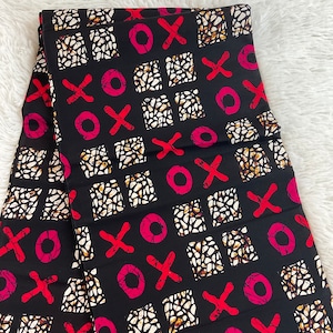 Könnte beinhalten: Schwarzer Stoff mit einem sich wiederholenden Muster aus roten X'en und rosa O's, zusammen mit weißen und braunen abstrakten Formen. Der Stoff ist gefaltet und zeigt die Wiederholung des Musters. Der Hintergrund ist eine flauschige weiße Oberfläche.