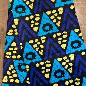 May include: A folded fabric with a vibrant geometric pattern. The design features large turquoise triangles and yellow circles against a black background, with blue zig-zag lines. The fabric appears to be cotton or a similar material.