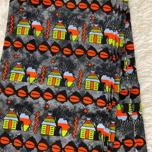 May include: A gray fabric with a repeating pattern of orange, yellow, and black African huts, trees, and sun symbols. The fabric is folded in a stack.