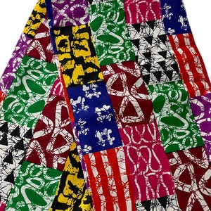 May include: A colorful patchwork fabric with a variety of geometric patterns. The fabric features squares of red, green, yellow, blue, and purple, each with unique designs in white and black. The fabric is folded, displaying its vibrant colors.