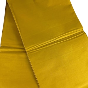 May include: A folded length of golden-yellow fabric, possibly silk or a similar material, with a subtle sheen. The fabric is smooth and appears to be of high quality, with visible folds and creases adding texture. The color is consistent throughout.