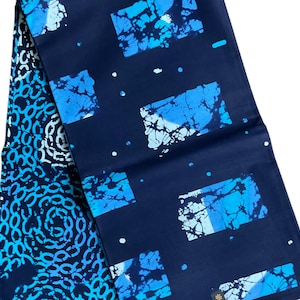 May include: A folded textile with a dark blue background and a repeating pattern of blue and white shapes. The design includes squares and circular patterns, creating a visually interesting contrast. The fabric appears to be cotton or a similar material.