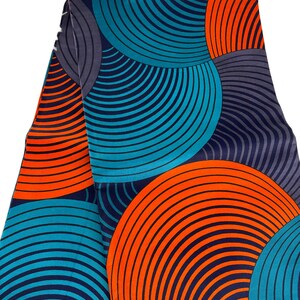 May include: Folded fabric with a vibrant circular pattern in teal, orange, and navy blue. The design features concentric arcs creating a dynamic, eye-catching visual. The fabric appears to be a medium weight, suitable for various sewing projects.