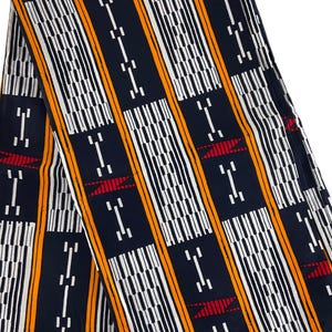 May include: Folded fabric with a repeating pattern of vertical stripes in white, orange, and black. The design includes geometric shapes and red accents, creating a bold and colorful textile.