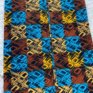 May include: A rectangular piece of fabric featuring a repeating grid pattern of alternating brown and bright blue squares. The surface is decorated with a dense, abstract print of yellow and black geometric shapes and textured, scratchy line work.