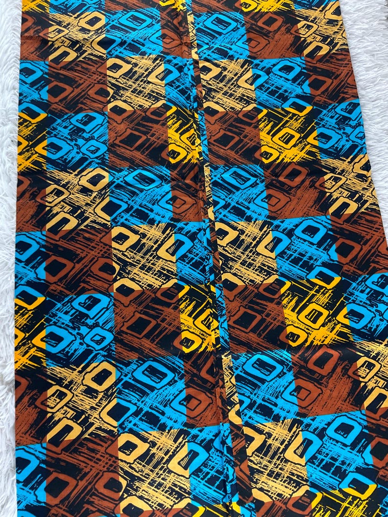 May include: A rectangular piece of fabric featuring a repeating grid pattern of alternating brown and bright blue squares. The surface is decorated with a dense, abstract print of yellow and black geometric shapes and textured, scratchy line work.