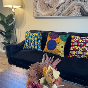 May include: Three square throw pillows featuring vibrant African-inspired patterns in blue, yellow, orange, green, red, and black, arranged on a black sofa. The pillows display geometric shapes, circular motifs, and intricate linear designs.