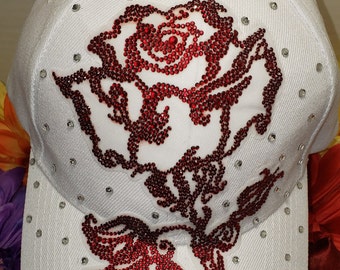 May include: White baseball cap with a red rhinestone rose design. The rose is detailed with a stem and leaves.