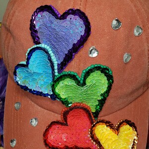 May include: A brown baseball cap with four sequined hearts in rainbow colors. The hearts are purple, blue, green, and red and orange. The cap has rhinestones on the front.