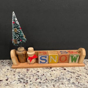 May include: A wooden Christmas decoration featuring a miniature tree, a snowman, and blocks spelling "SNOW." The tree is green with red berries and pinecones. The snowman has a red ribbon. The blocks are painted with colorful letters.