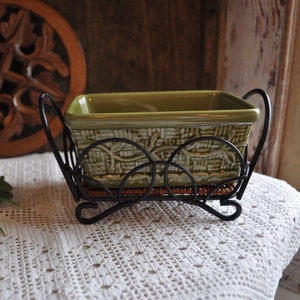 May include: A green ceramic rectangular dish with a woven pattern, set in a black wrought iron holder. The holder has a decorative design and sits on a woven base. The dish is likely for serving or display.
