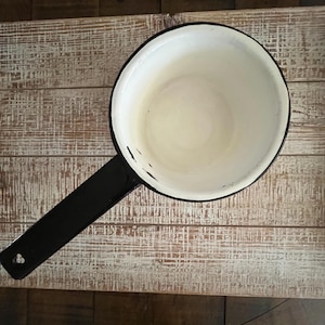 May include: A vintage enamel saucepan with a black handle and rim. The interior is white, and the exterior is black. The saucepan is set against a weathered, light-colored wooden surface, suggesting a rustic or farmhouse aesthetic.