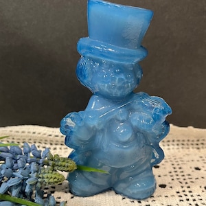 May include: A light blue figurine of a character wearing a top hat and holding a bouquet. The figurine has a glossy finish and is set against a neutral background. The character's features are detailed, and the overall aesthetic is whimsical.