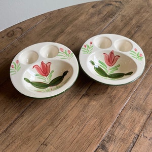 May include: Two white ceramic egg cups with a green rim and a floral design. The design features a pink tulip with green leaves and small pink flowers.