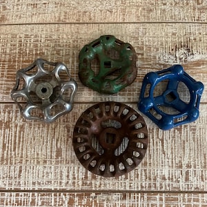 May include: Four vintage water valve handles in silver, green, brown, and blue. The handles are made of metal and have various spoke and circular designs. The handles are arranged on a weathered, light-colored wooden surface.