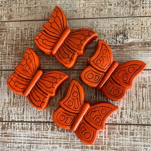 May include: Four orange butterfly-shaped decorative objects with dark brown etched details. The butterflies are arranged on a light brown, textured wooden surface. The wings have a detailed pattern.