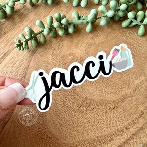 K&ouml;nnte beinhalten: Ein wei&szlig;er Aufkleber mit dem Namen "jacci" in schwarzer Kursivschrift. Der Aufkleber zeigt eine Schere, eine Haarb&uuml;rste und eine Flasche Haarf&auml;rbemittel, was auf ein Thema f&uuml;r Friseure oder Salons hindeutet.
