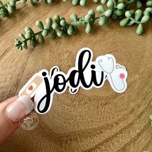 K&ouml;nnte beinhalten: Ein schwarz-wei&szlig;er Aufkleber mit dem Namen "Jodi" in Kursivschrift. Der Aufkleber zeigt au&szlig;erdem ein kleines rotes Herz, ein wei&szlig;es Pflaster und ein Stethoskop.
