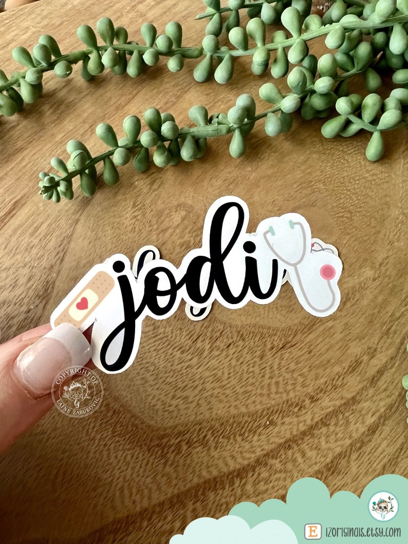 K&ouml;nnte beinhalten: Ein schwarz-wei&szlig;er Aufkleber mit dem Namen "Jodi" in Kursivschrift. Der Aufkleber zeigt au&szlig;erdem ein kleines rotes Herz, ein wei&szlig;es Pflaster und ein Stethoskop.