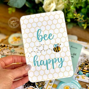 May include: A white card with a honeycomb pattern and the text "bee happy" in blue. A cartoon bee with black and yellow stripes is flying above the text.