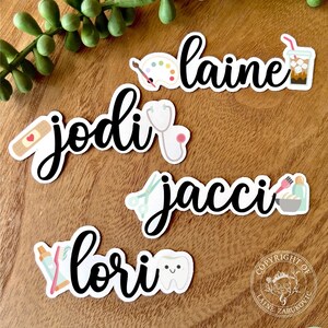 K&ouml;nnte beinhalten: F&uuml;nf personalisierte Namenaufkleber mit verschiedenen Symbolen. Die Namen sind "JODI", "LAINE", "JACCI", "LORI" und ein Copyright-Symbol mit dem Text "COPYRIGHT OF LAINE ZABUKOVIC".