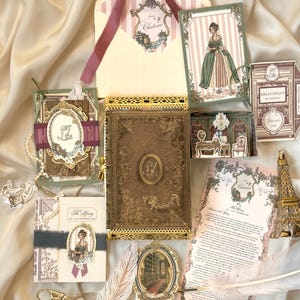 May include: A collection of stationery items including a central brown book with gold filigree, an envelope with a mauve ribbon, illustrated cards featuring a person in a green dress, a small gold pair of scissors, a white feather quill, and a miniature Eiffel Tower ornament.