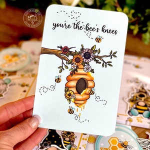 May include: A white card with the text "you're the bee's knees" in black lettering. The card features a cartoon illustration of a beehive with bees and flowers.