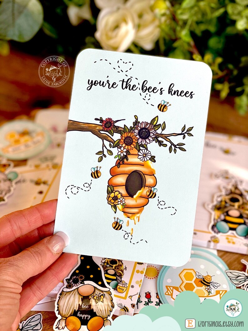 May include: A white card with the text "you're the bee's knees" in black lettering. The card features a cartoon illustration of a beehive with bees and flowers.