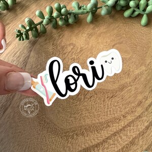 K&ouml;nnte beinhalten: Ein wei&szlig;er Aufkleber mit dem Namen "Lori" in schwarzer Kursivschrift. Neben dem Namen befinden sich ein Cartoon-Zahn mit einem fr&ouml;hlichen Gesicht und eine rosa-blaue Zahnb&uuml;rste. Der Aufkleber befindet sich auf einer Holzoberfl&auml;che.