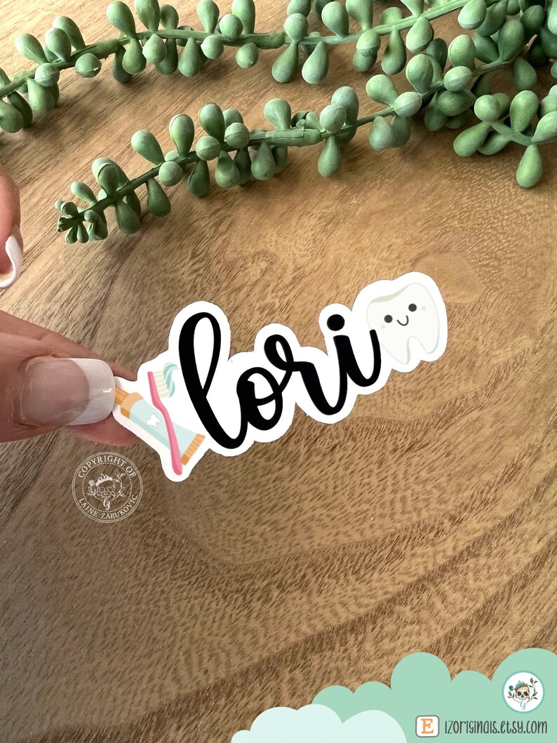 K&ouml;nnte beinhalten: Ein wei&szlig;er Aufkleber mit dem Namen "Lori" in schwarzer Kursivschrift. Neben dem Namen befinden sich ein Cartoon-Zahn mit einem fr&ouml;hlichen Gesicht und eine rosa-blaue Zahnb&uuml;rste. Der Aufkleber befindet sich auf einer Holzoberfl&auml;che.