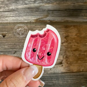以下が含まれることがあります： ピンク色の棒アイスのステッカー。一部が噛み取られています。ステッカーには笑顔の顔とキラキラしたアクセントがあります。木製の背景にあります。