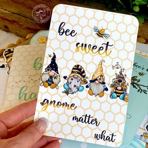 May include: A white card with a honeycomb pattern and the text "bee sweet gnome matter what". There are four gnomes with bee wings and a bee on the card.