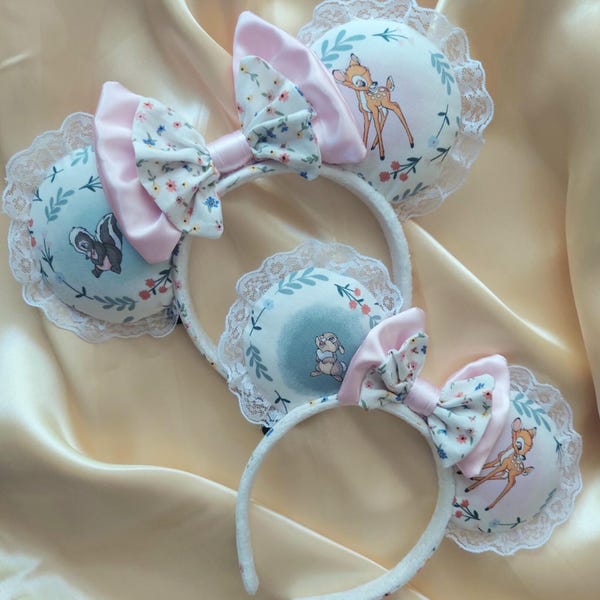 May include: Three white headbands with plush ears featuring woodland creature designs. Each headband has a pink bow and floral accents. The ears are round with lace trim and illustrations of a skunk, a bunny, and a fawn.