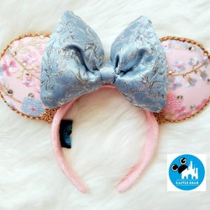 May include: A pair of mouse ears with a light blue bow and pink ears. The ears are decorated with floral embroidery and gold trim. The headband is pink and has a tag that says "Castle Gear".