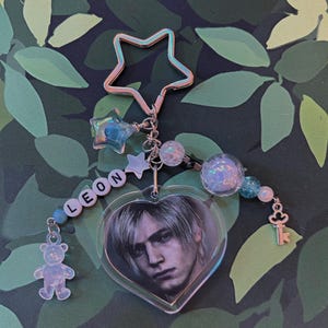 Pu&ograve; includere: Un portachiavi con un ciondolo a forma di cuore con un ritratto, il nome "LEON" in perline di lettere e abbellimenti a forma di stella e perline. Include un piccolo ciondolo a forma di orso e un ciondolo a forma di chiave.