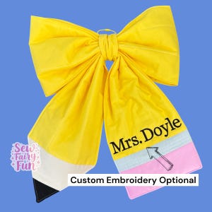 May include: A large yellow fabric hair bow shaped like a pencil, featuring a black tip, a white band, and a pink eraser section. The text "Mrs. Doyle" is embroidered in black on the yellow portion, accompanied by a small black arrow graphic.