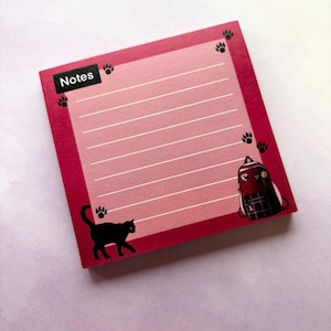 May include: Pink and red notepad with lined paper and a black cat and a red backpack with a plaid pattern on the front. The notepad has the word "Notes" printed on the top.