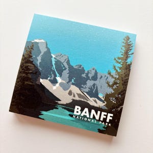 Puede incluir: Una impresión cuadrada con un paisaje montañoso con un lago y árboles. La impresión tiene un cielo azul y el texto "BANFF NATIONAL PARK" en blanco. La obra de arte es de estilo póster de viaje vintage.