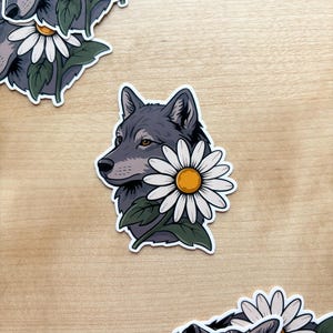 May include: A sticker featuring a gray wolf with a white daisy flower. The wolf is in profile, with detailed fur and amber eyes. The daisy has white petals and a yellow center, with green leaves. Additional stickers are scattered around.