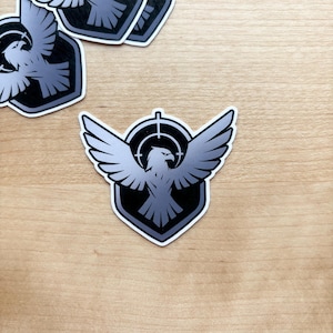 May include: A collection of stickers featuring a stylized eagle with outstretched wings, centered within a target reticle. The eagle and target are in shades of gray and black, set against a white border and a black shield shape. The stickers are on a light brown surface.