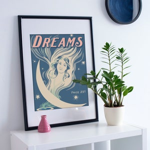May include: A vintage-style poster with the word "DREAMS" in pink lettering. The poster features a woman with long, flowing hair, surrounded by stars and a crescent moon. The poster is framed in black and hanging on a white wall.