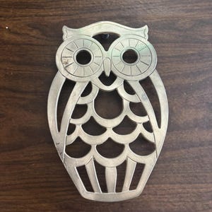 May include: A brass-colored owl-shaped trivet with a cutout design. The trivet features large circular eyes, a scalloped chest, and a small loop at the top for hanging. The owl is set against a dark brown wooden surface.