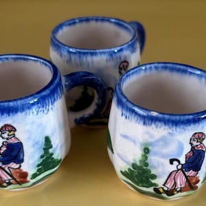 May include: Three white ceramic mugs with blue trim and hand-painted scenes of people in a landscape. Each mug has a different person and a different landscape.
