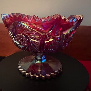 May include: A large, iridescent, cranberry glass bowl with a scalloped edge and a pedestal base. The bowl has a decorative, floral pattern.