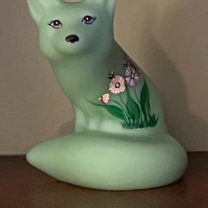 May include: A light green glass figurine of a fox with painted details. The fox has a long, curled tail and is sitting with its head tilted slightly to the side. The fox has painted eyes, a black nose, and a floral design on its back.